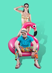A black haired women in sunglasses and a gold bikini behind an inflatable flamingo that is sat on by a guy in a fedora, aviators, rainbow pants and a blue shirt.