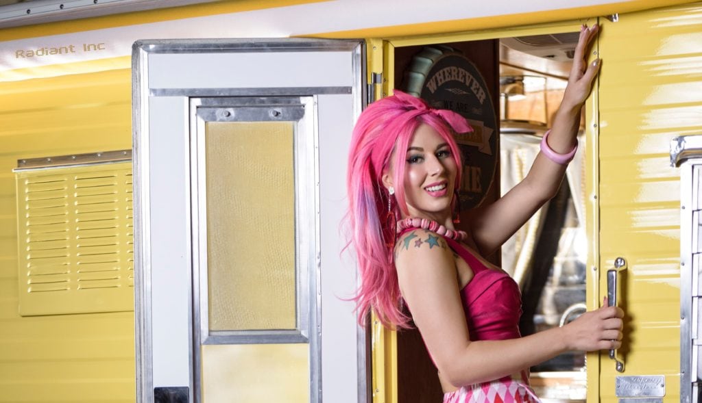 Pink hair girl in doorway of a yellow tanning room with a pink top, pink checkered skirt, a pink bracelet and a star shoulder tattoo